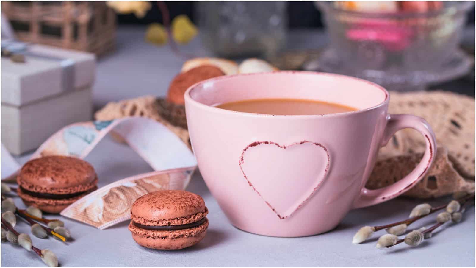 A pink ceramic mug with a heart imprint is filled with coffee or tea. Two brown macarons are placed next to the mug. Decorative items, including a lace cloth, a ribbon, and fuzzy branches, are in the background, along with a wrapped gift box.
