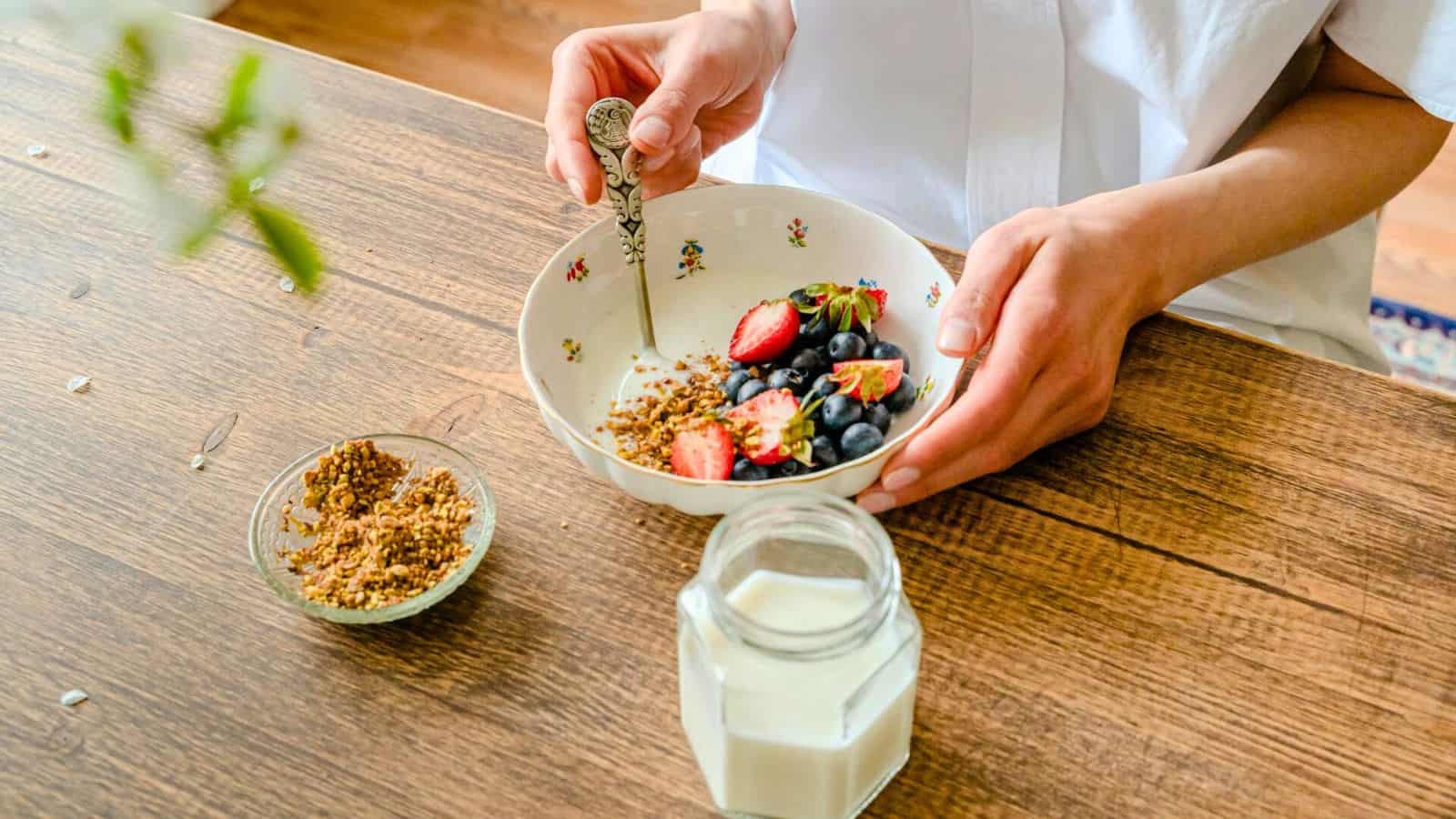 A person seated at a wooden table is holding a spoon and a bowl containing granola, strawberries, blueberries, and yogurt. A small dish filled with granola and a jar of milk are also on the table. The person is wearing a white shirt.