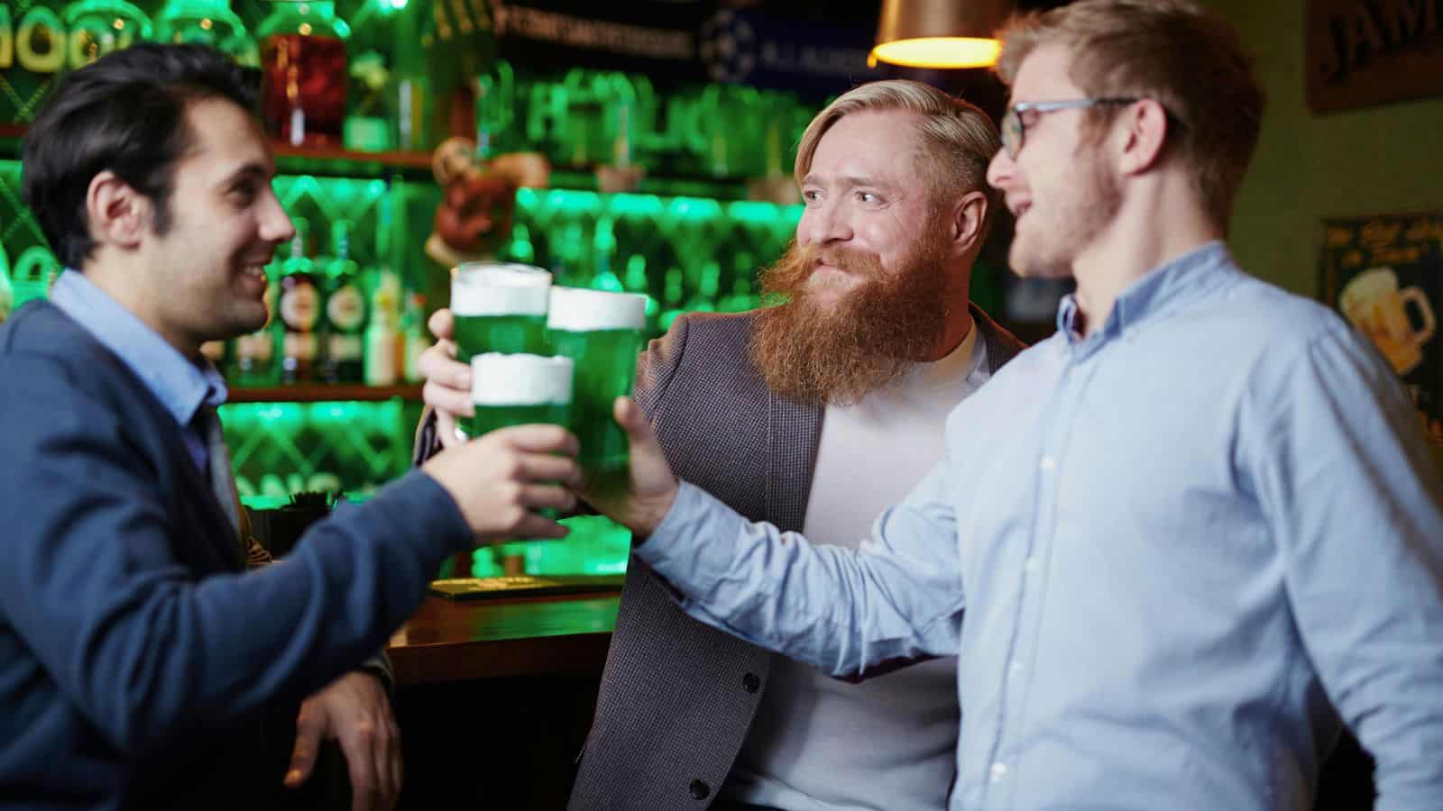Three men are seen toasting with fun green drinks in a bar. They are all smiling and wearing casual clothing. The bar has green lighting, and shelves with various bottles are visible in the background.