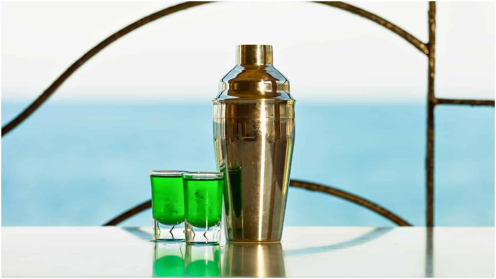 A metal cocktail shaker and two small glasses filled with green liquid are placed on a white surface. The background shows a blurred view of the ocean with metal railing details, making it look like an idyllic setting despite featuring types of alcohol you shouldn't be drinking.