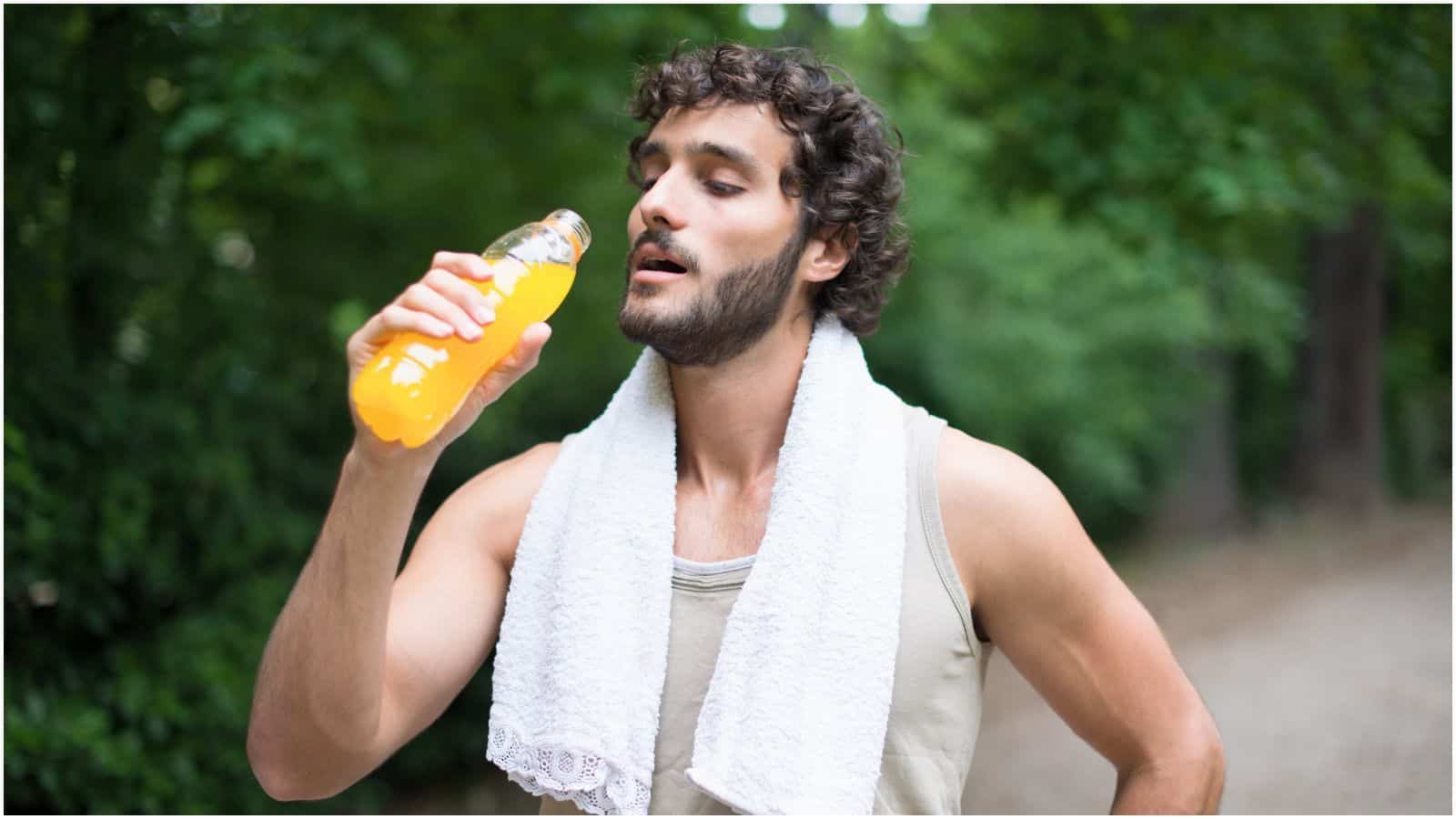 A person with curly hair and a beard is outdoors, holding a bottle of orange sports drink&mdash;clearly no interest in the types of alcohol you shouldn't be drinking. They are wearing a beige tank top and have a white towel draped around their neck. Trees and greenery can be seen in the background.