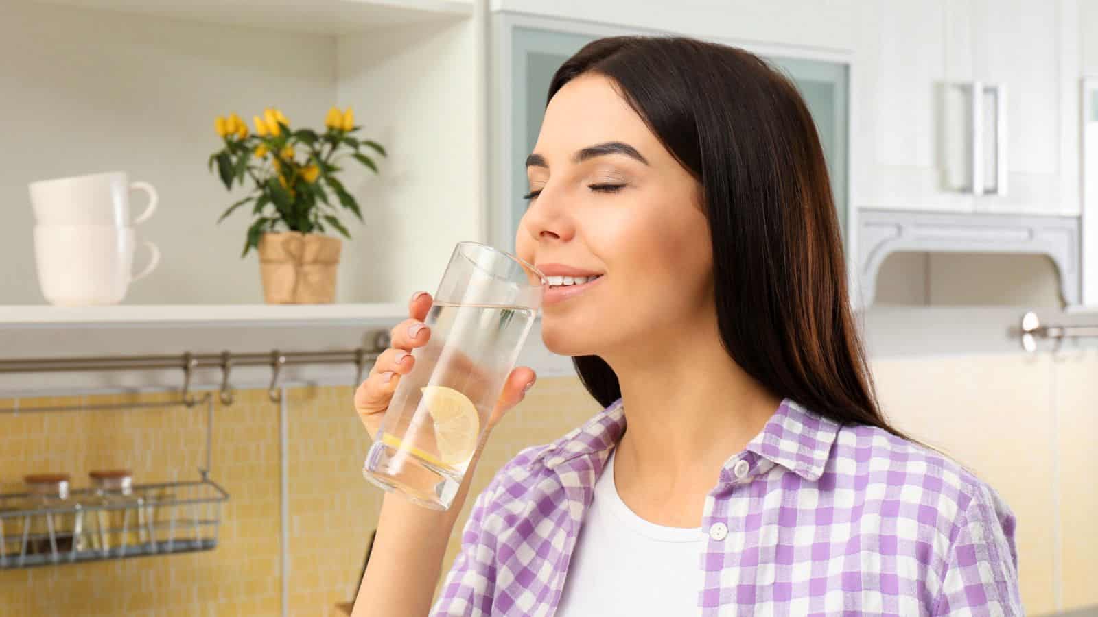 A woman with long brown hair is standing in a kitchen, smiling with her eyes closed while holding a glass of lemon water before bed. She is wearing a purple and white checkered shirt. There are kitchen cabinets, a counter, and a potted plant with yellow flowers in the background.