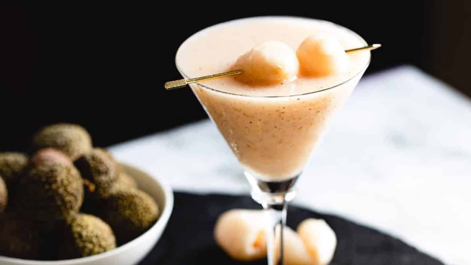 A martini glass filled with a light pink smoothie is garnished with two round fruits on a skewer. A white bowl filled with brown, round fruits is placed next to the glass, adhering to alcohol rules. The scene is set on a black and white surface with a dark background.
