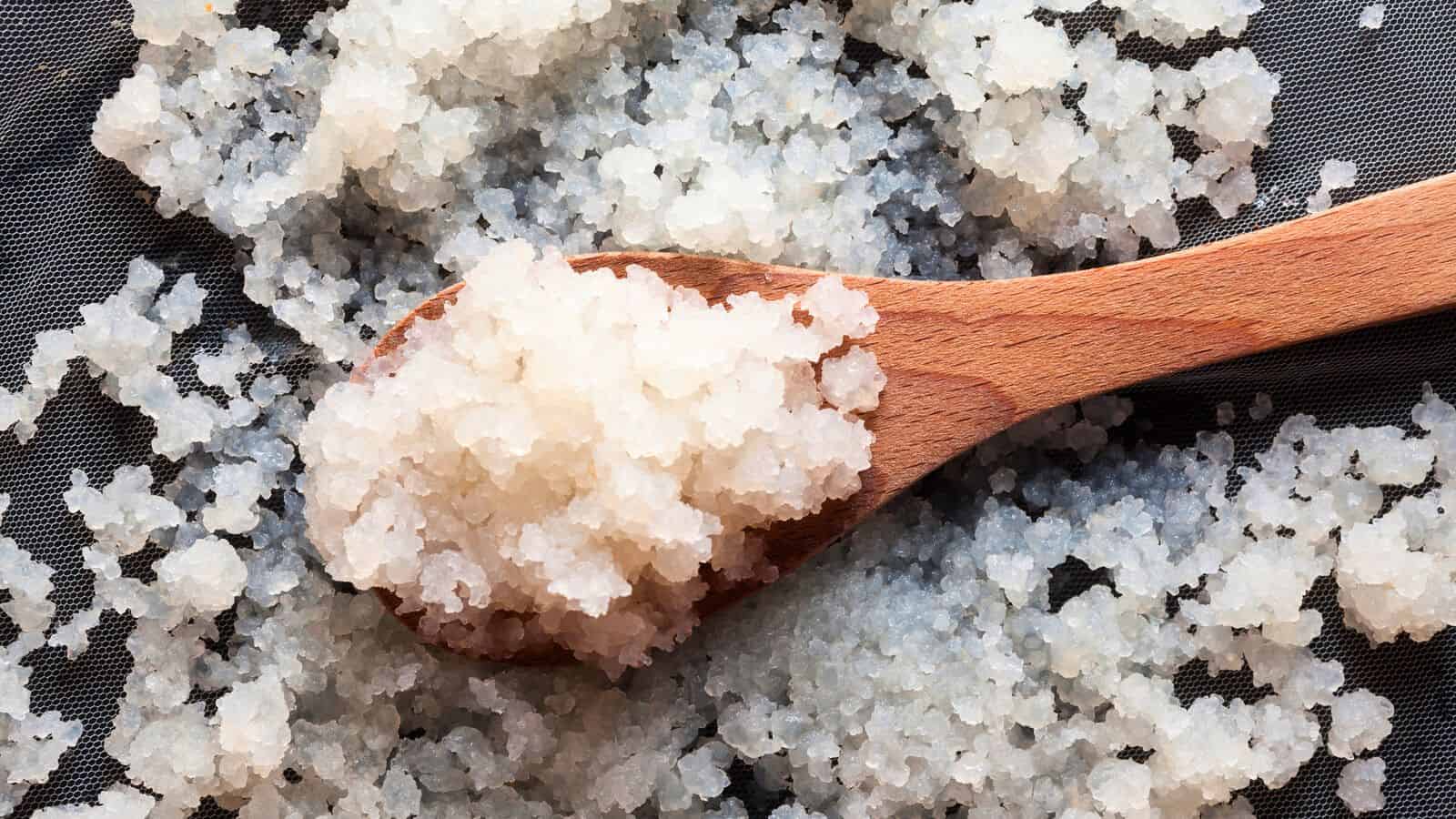 A close-up image of a wooden spoon holding a cluster of kefir. More of the same grains are scattered around the spoon on a dark, textured surface.