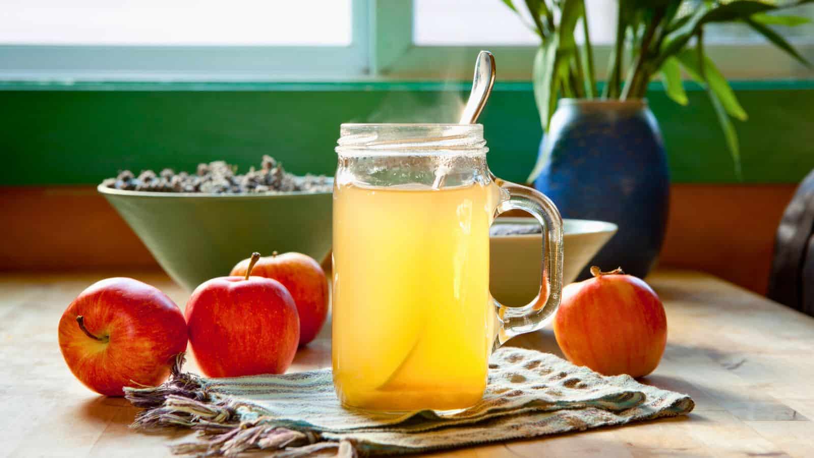 A steamy glass mug of hot apple cider sits on a wooden table, surrounded by three red apples and a green and white cloth. Behind them, there's a green bowl filled with dried lavender and a blue vase with leafy green plants. A window provides natural light.