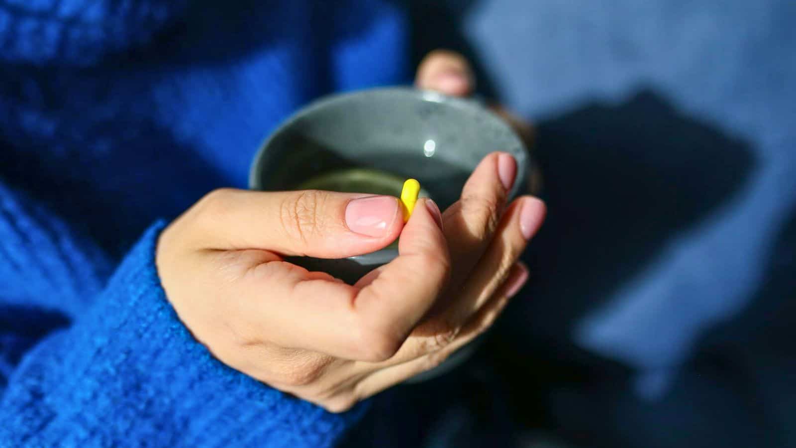 Close-up of a person holding a small yellow capsule in one hand and a gray cup in the other, adhering to strict alcohol rules. The individual is wearing a blue sweater, with light pink nail polish on their nails. The background is blurred and dark.