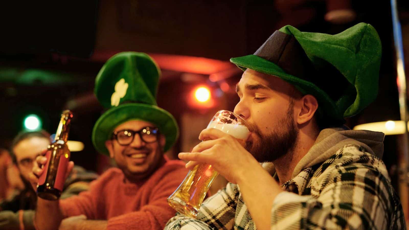 Two individuals at a bar celebrating, both wearing large green hats. One person is drinking from a beer glass while the other is holding a beer bottle and smiling. Despite the festive mood, they are mindful of alcohol rules in the dimly lit bar with colorful lights in the background.