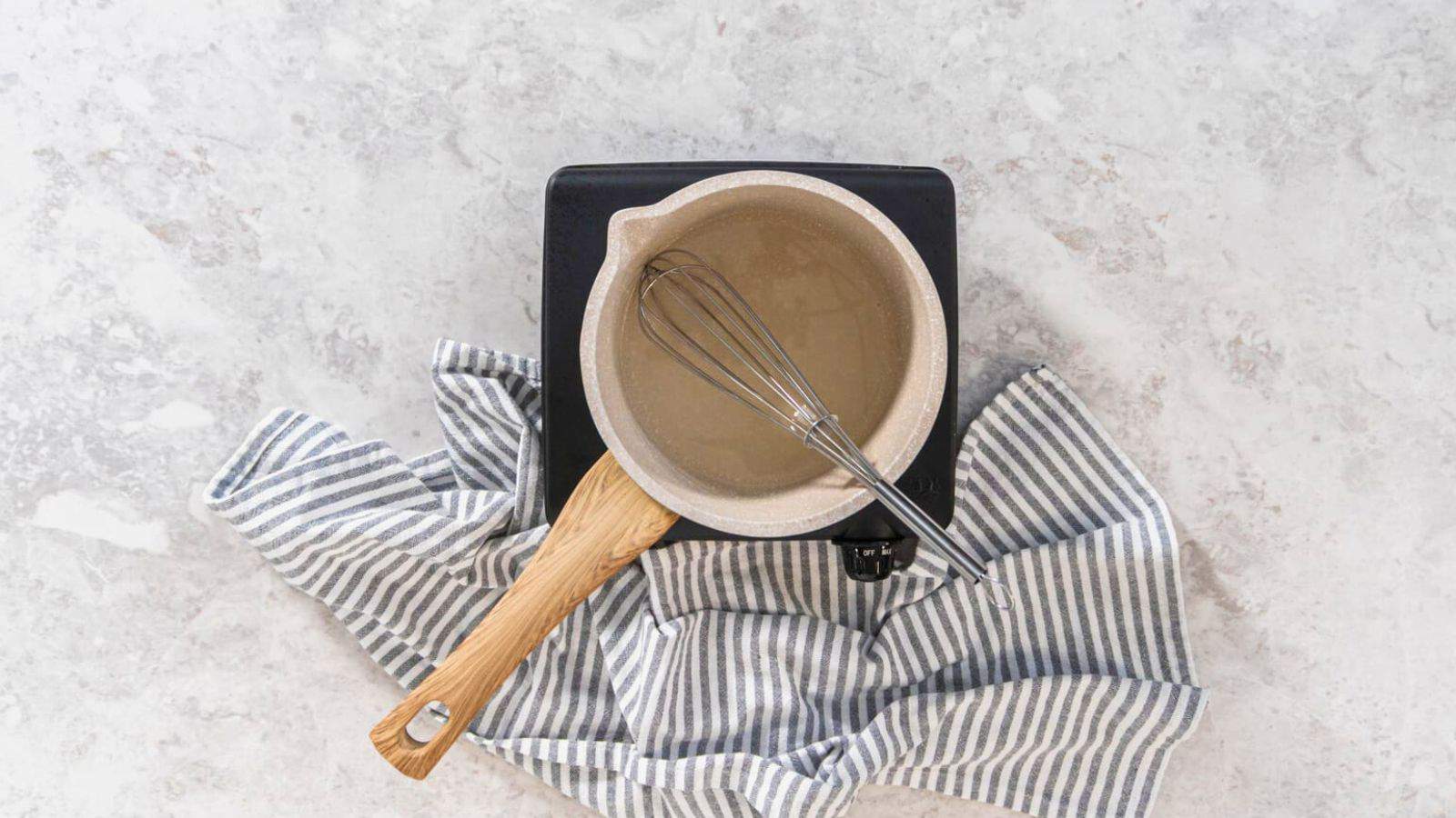 Overhead view of a small pot with a wooden handle on a single burner hot plate. A whisk is inside the pot, which is filled with light-colored simple syrup. Below the hot plate, there is a blue and white striped cloth spread out on a light, textured surface&mdash;everything you need to know about simple syrup in one image.