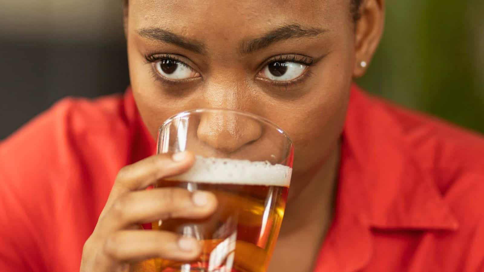 A person wearing a red shirt is holding a glass of beer close to their mouth, appearing to take a sip. The beer has a foamy head and the person's expression is focused and calm, mindful of alcohol rules. The background is slightly blurred.