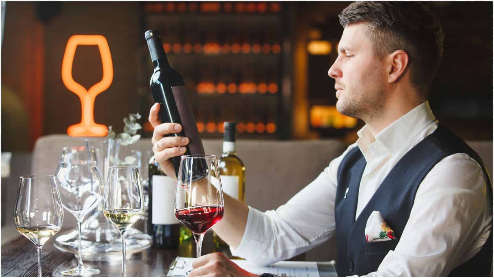 A man in a white shirt and dark vest holds a bottle of wine, carefully inspecting the label. In front of him are multiple wine glasses, some filled with red and white wine. The background showcases an array of wine bottles and a neon glass sign&mdash;an ode to mastering alcohol rules.