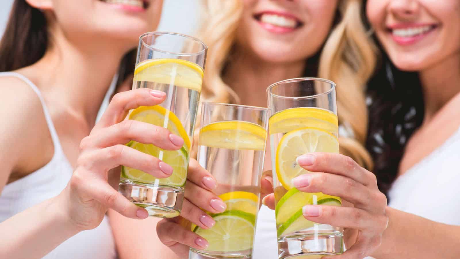 Three people are holding glasses of water with lemon and lime slices, toasting together. They are smiling and wearing white tops. The focus is on the glasses in the foreground, highlighting the refreshing appeal of lemon water before bed.
