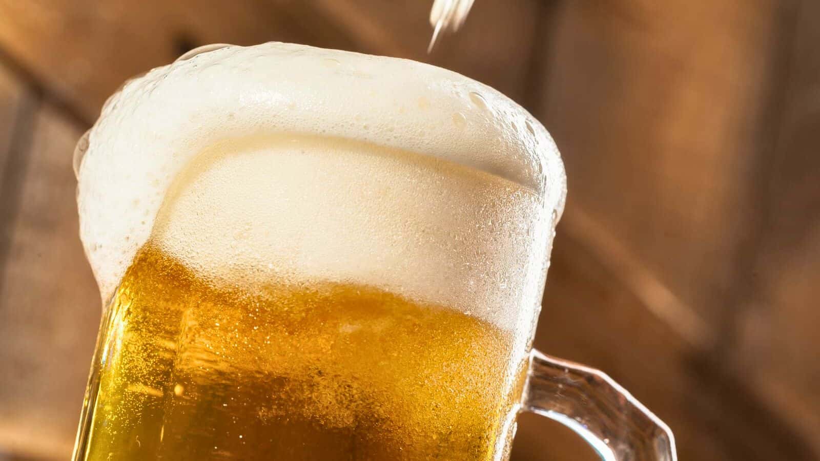 Close-up of a frothy beer being poured into a large glass mug. The mug is tilted slightly, and the beer has a thick white foam on top. The background is blurred with warm, wooden tones, highlighting the focus on the beer, one of those tasty alcoholic drinks that are bad for your teeth.