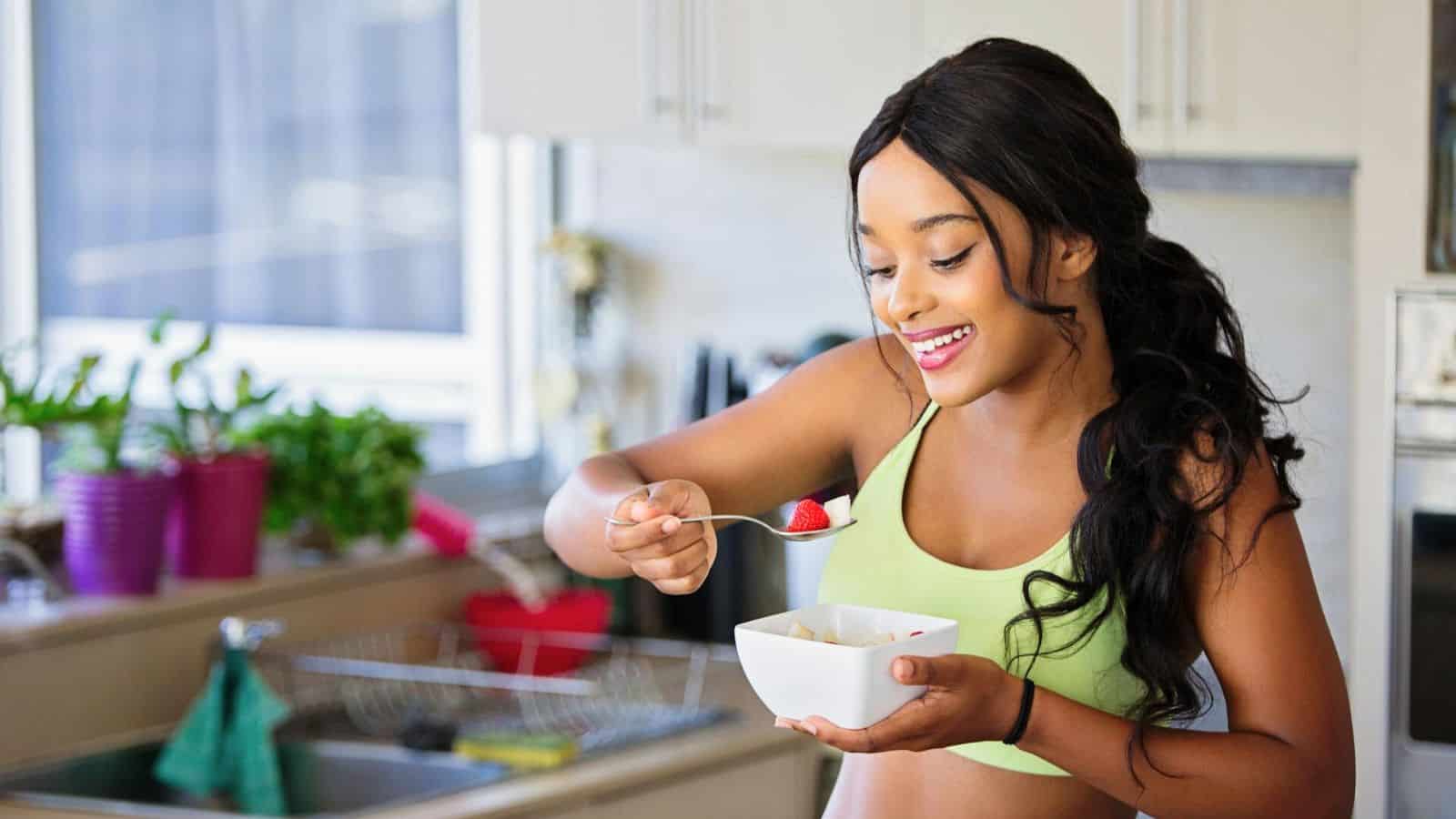 A woman is standing in a kitchen, smiling as she holds a spoon with a piece of fruit and a bowl of food. She is wearing a green sports bra and has long dark hair. Potted plants and kitchen utensils are visible in the background, suggesting her healthy lifestyle after deciding to stop drinking alcohol.