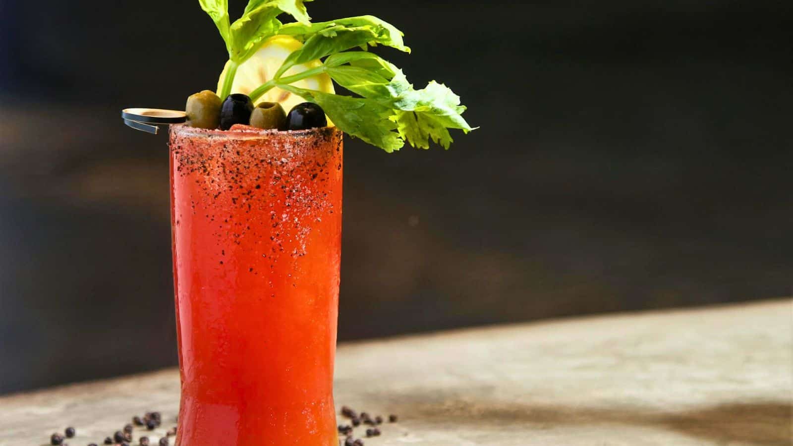 A tall glass of Bloody Mary garnished with olives, a lemon slice, a celery stalk, and pepper around the rim. The drink is red, and the background is blurred. Black peppercorns are scattered on the surface around the base of the glass&mdash;one of those alcoholic drinks that are bad for your teeth.
