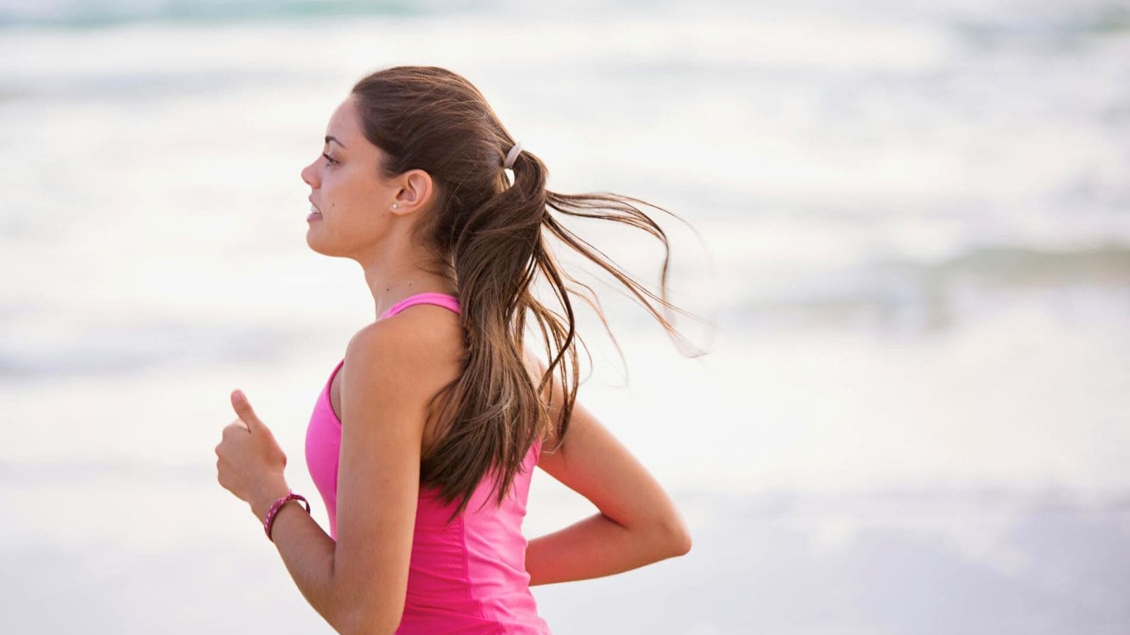 A person with long hair tied in a ponytail is running on a beach, enjoying the fresh sea air. They are wearing a sleeveless pink top. The background features a sandy shore with waves in the distance.