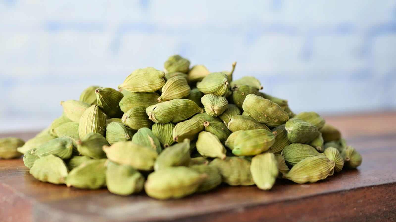A close-up image shows a pile of green cardamom pods placed on a wooden surface. The background is softly blurred with a blue shade, drawing focus to the spices for coffee. The cardamom pods are small and oval, with a distinct ridged texture.