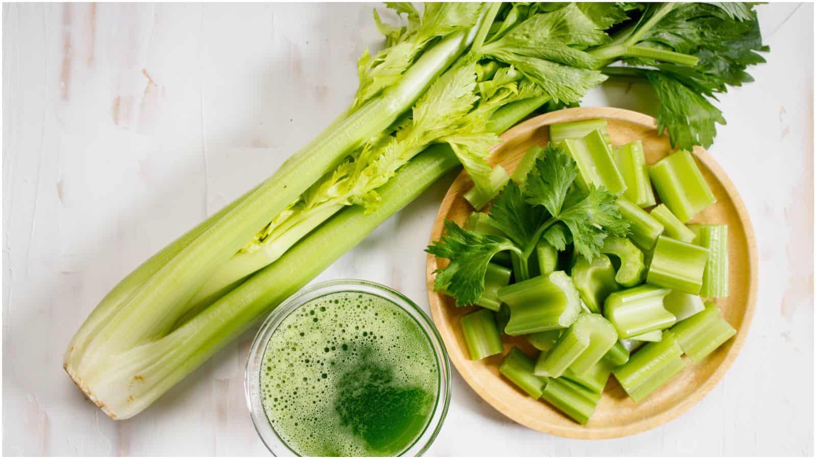 A whole celery stalk, a glass of green celery juice&mdash;often recommended as a drink for gout&mdash;and a bowl filled with chopped celery pieces are arranged on a light-colored surface. The celery stalk has long green leaves, and the juice appears freshly made with a frothy top.