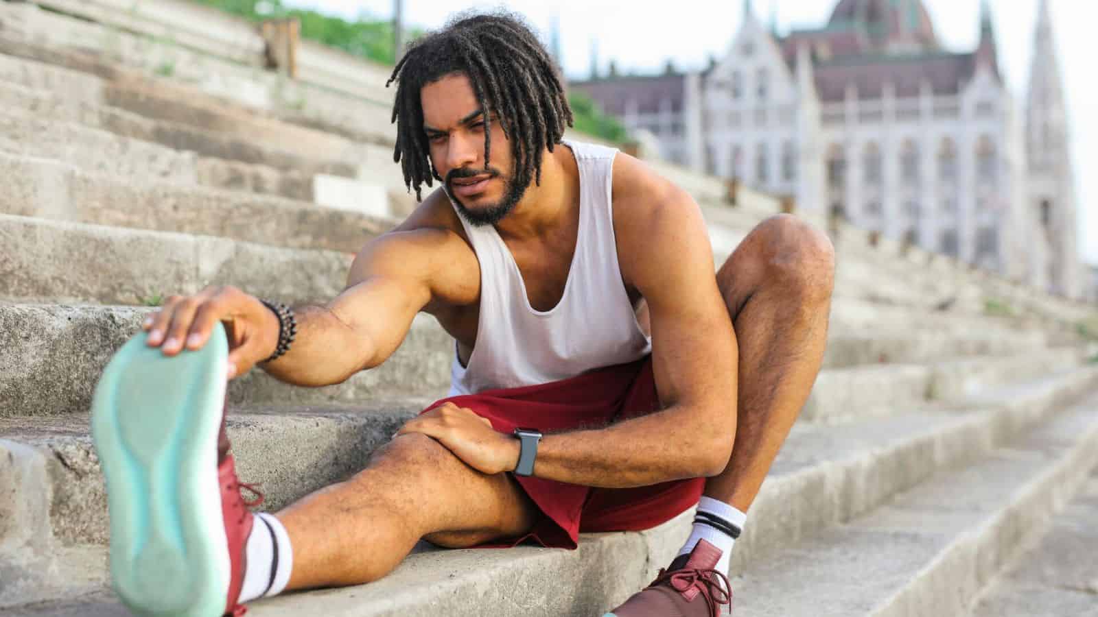 A man wearing a white tank top, red shorts, and light blue shoes is stretching on outdoor stairs. He is sitting with one leg extended and reaching for his foot. In the background, a large, ornate building can be seen as he contemplates the best drinks for gout to stay healthy.