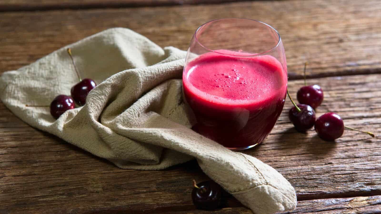 A glass of red juice sits on a wooden surface next to a light-colored cloth. Surrounding the cloth and glass, perfect as drinks for gout relief, are several fresh cherries with stems. The scene is set with a rustic aesthetic.