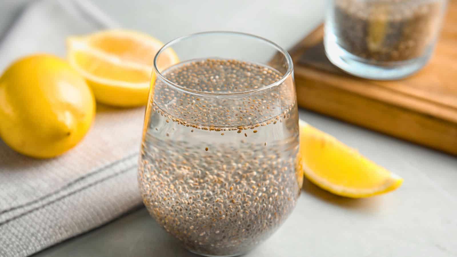 A glass filled with water and chia seeds sits on a table. Beside the glass are two lemon halves and one lemon wedge placed on a cloth and wooden board. Another glass with a similar mixture is partially visible in the background.