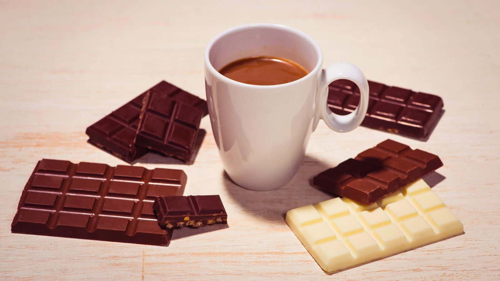 A white mug filled with coffee is placed on a light-colored surface. Surrounding the mug are various chocolate bars, including dark chocolate, milk chocolate, and white chocolate, some partially broken. This cozy setup might make you rethink that list of sugary drinks in favor of simpler pleasures.