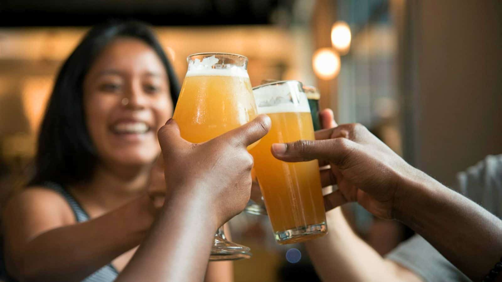 Three individuals clink their beer glasses together in a toast, celebrating in a casual indoor setting. One person, slightly blurred and smiling, is visible in the background. The beer glasses are filled with light amber beer, adhering to all alcohol rules for responsible enjoyment.