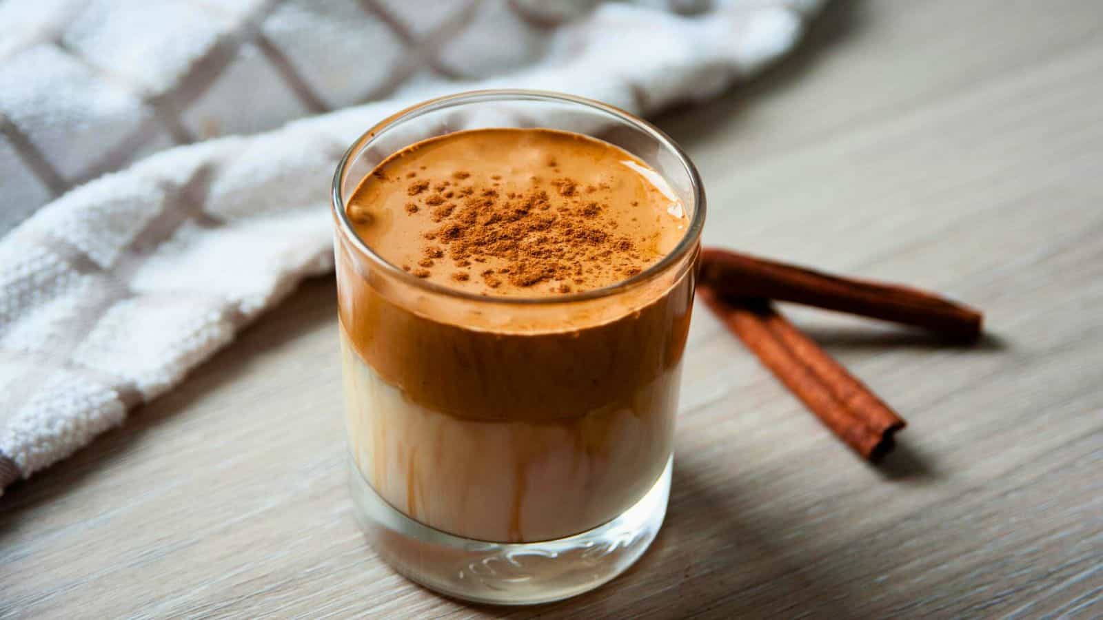A glass of dalgona coffee on a wooden surface. The drink is layered, with frothy whipped coffee on top and milk at the bottom, garnished with a sprinkle of cinnamon. There is a textured white towel in the background, along with two cinnamon sticks&mdash;ideal spices for coffee lovers.