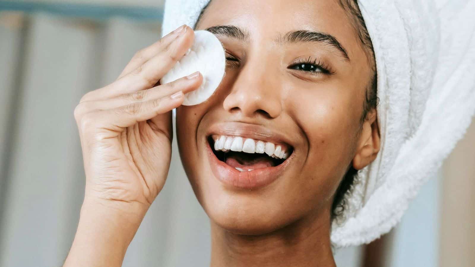 A person with a white towel wrapped around their head is smiling, holding a cotton pad to their face, and radiating the joyful benefits of choosing to stop drinking alcohol. The background is slightly blurred.
