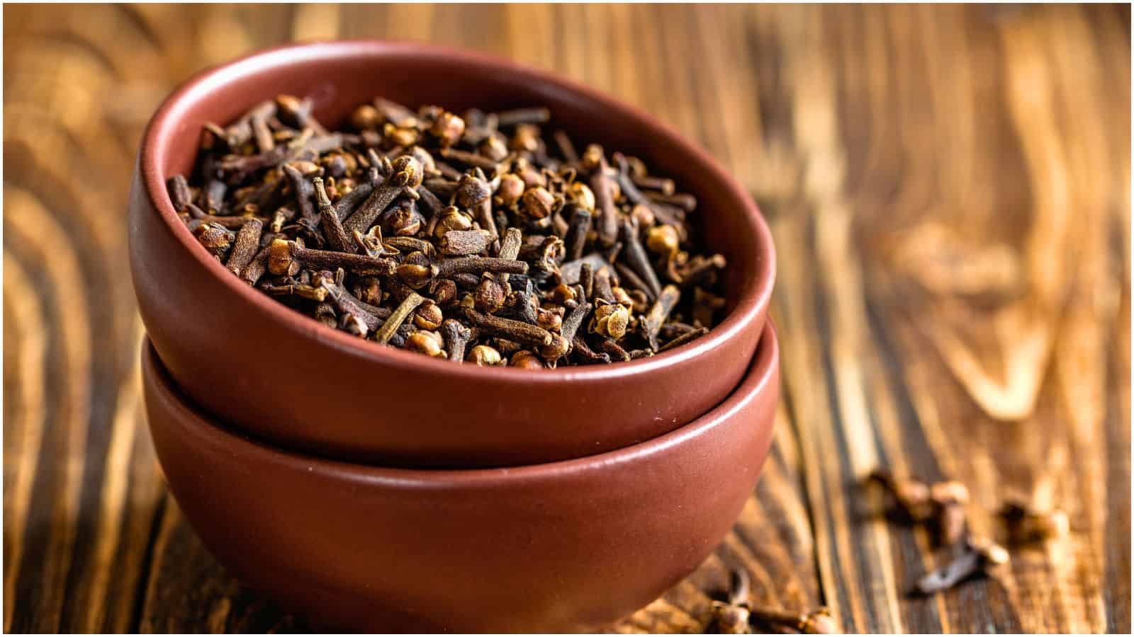 A bowl filled with dried cloves, often used as spices for coffee, sits atop another empty bowl. The bowls are placed on a wooden surface, and a few loose cloves are scattered around them. The image showcases the cloves prominently against the wooden background.