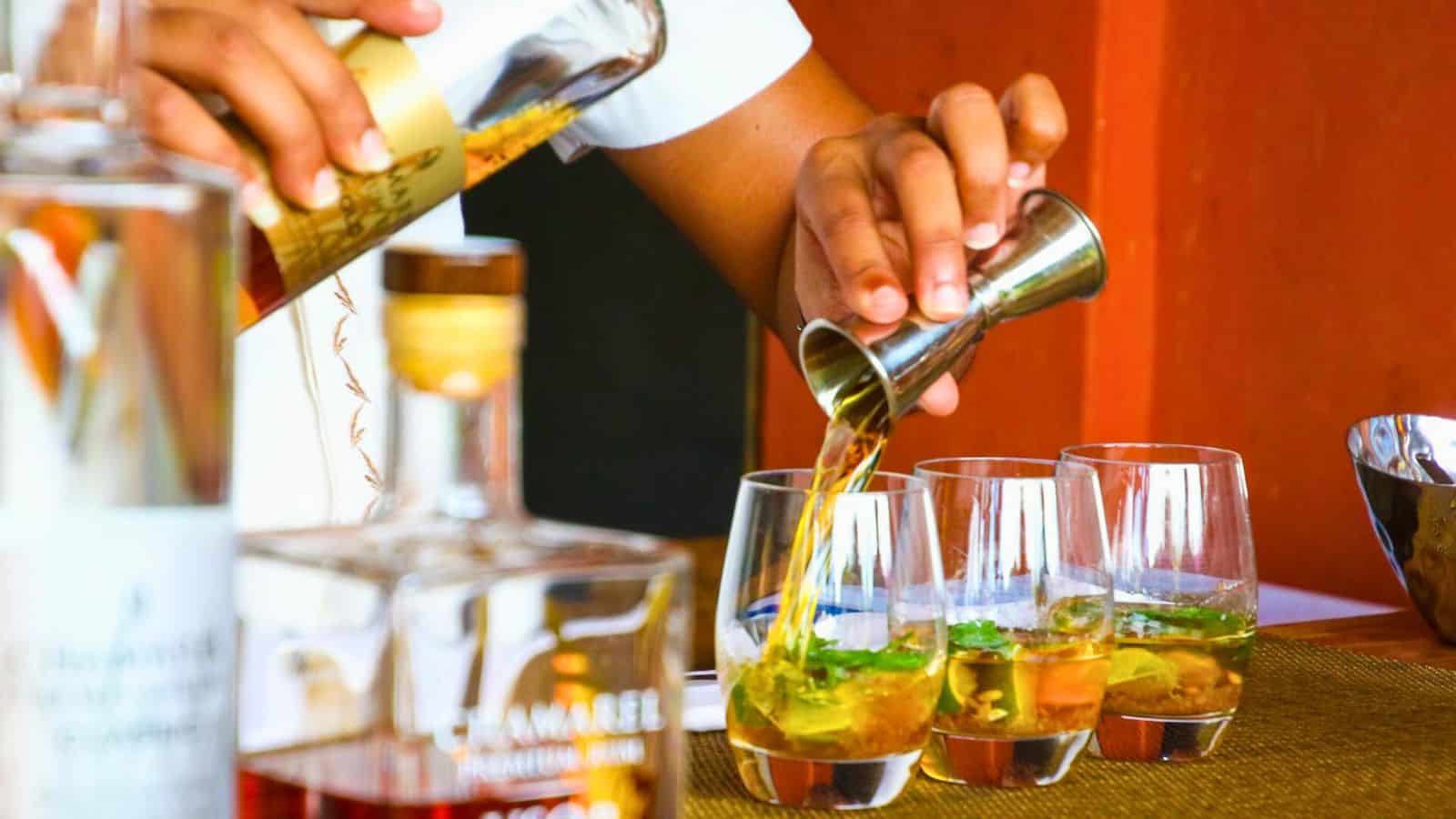 A person pours a liquid from a jigger into a glass on a countertop. Several glasses containing ice and garnishes are lined up on the counter, resembling a vibrant list of sugary drinks. Bottles of various spirits are placed nearby.