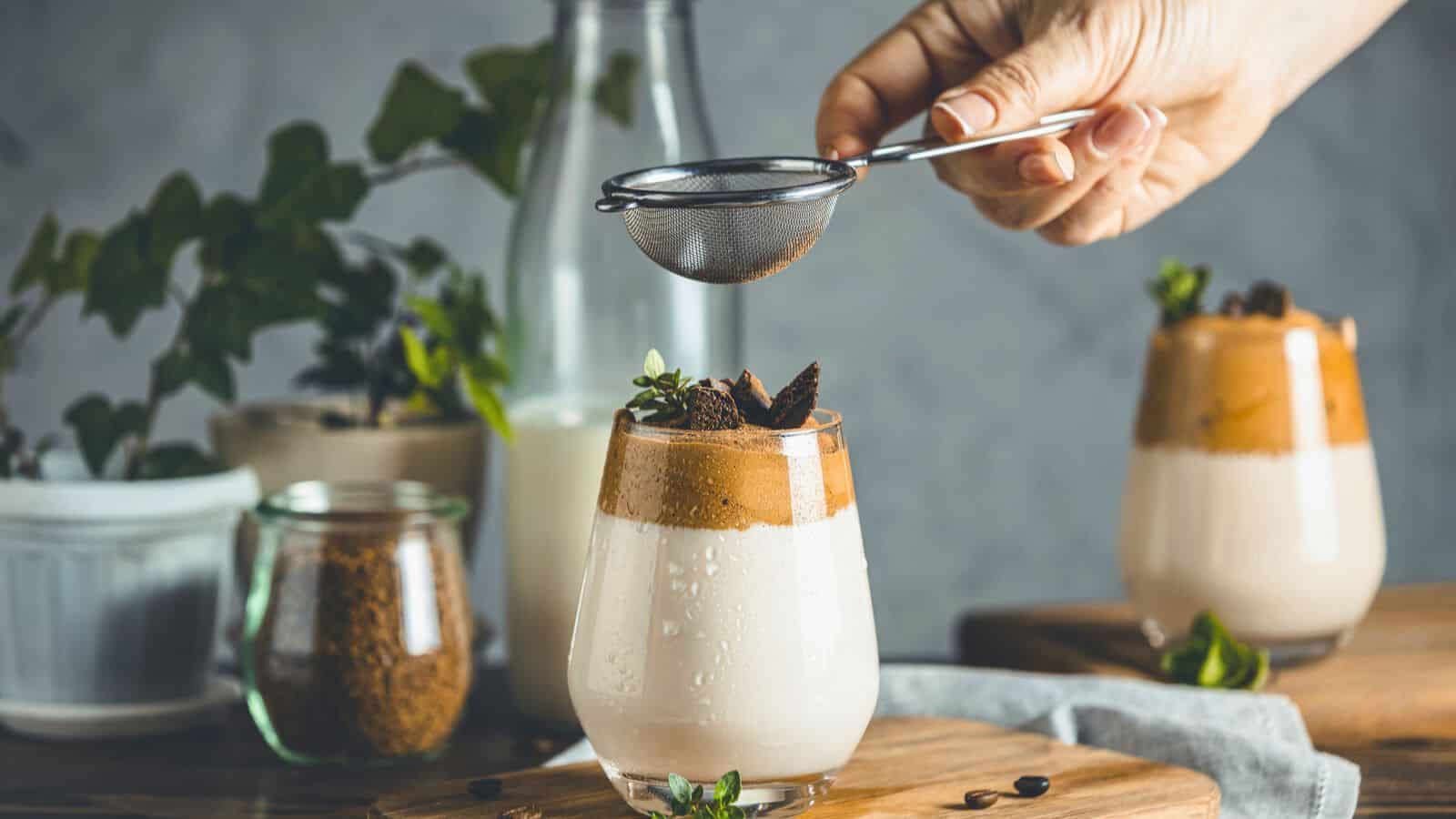 A hand holds a small strainer, sprinkling cocoa powder and spices for coffee onto a whipped coffee drink in a clear glass. Another identical drink is visible in the background. The scene includes various jars and a milk bottle, all arranged on a wooden board with a gray backdrop.