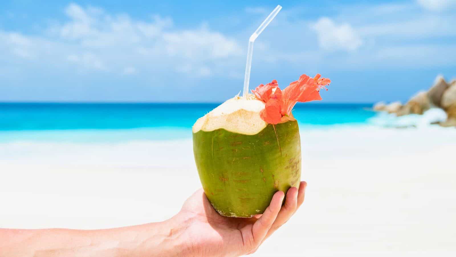 A hand is holding a fresh green coconut with a straw and a decorative tropical flower on a sunny beach, offering a refreshing escape from the usual list of sugary drinks. The background features a clear blue sky and turquoise ocean waves.