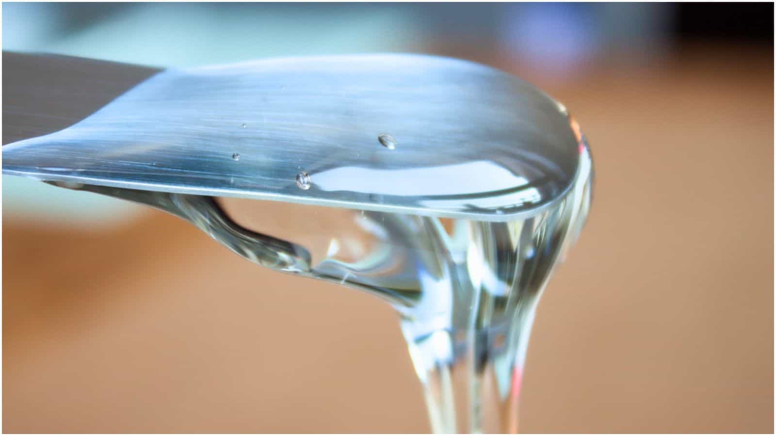 Close-up image of a metallic utensil with clear, viscous liquid drizzling off its edge. The shiny, reflective liquid flows downward, creating a glossy surface. Everything you need to know about simple syrup is captured in this mesmerizing instant. Background is blurred and out of focus.
