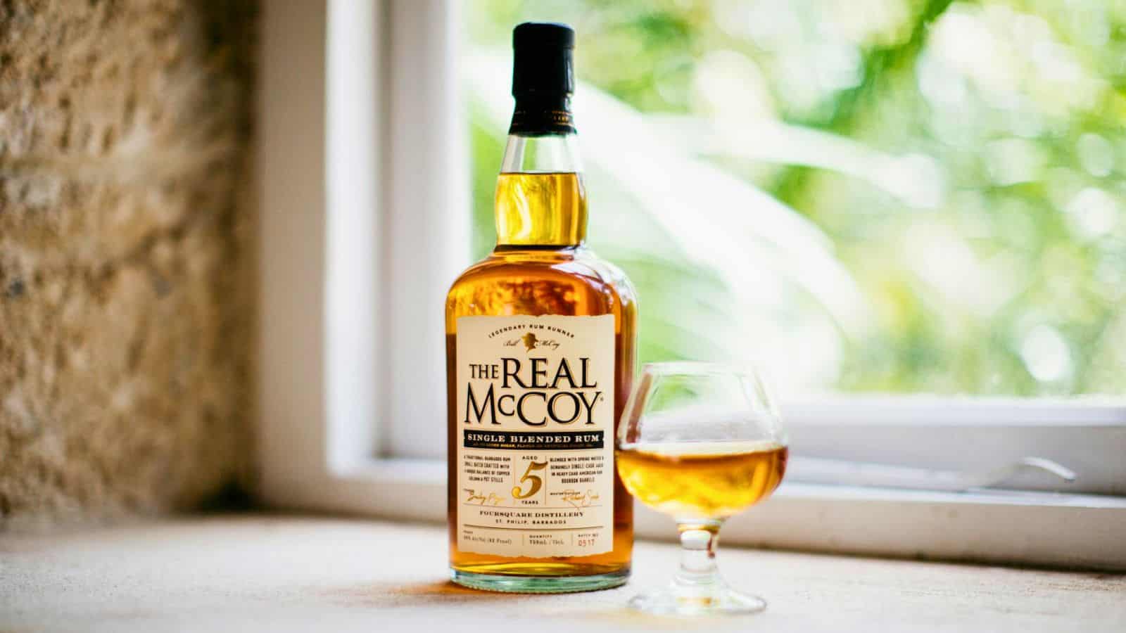 A bottle of The Real McCoy 5-year-old single blended rum stands next to a filled glass in front of a window with a blurry green outdoor background. Unlike some types of alcohol you shouldn't be drinking, this rum boasts a refined label in white with black and gold text, complementing the amber liquid in the glass.
