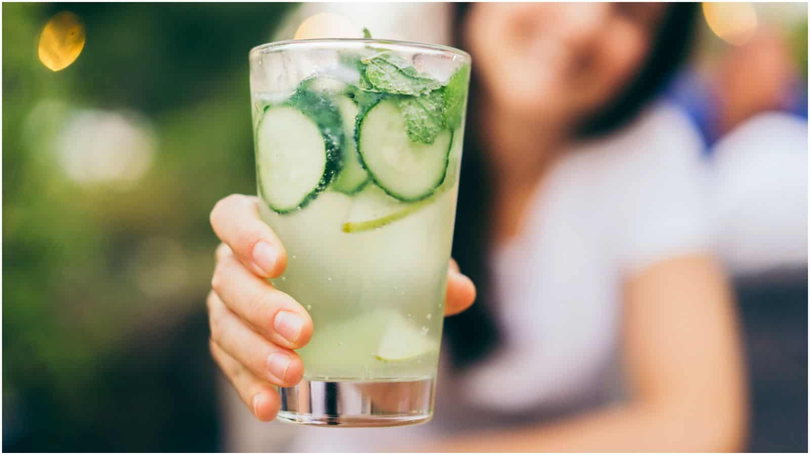 A person holding a clear glass of a greenish beverage garnished with cucumber slices, mint leaves, and lime wedges. The background is blurred, focusing on the drink and the hand holding it. This refreshing choice could be among recommended drinks for gout sufferers. The person is wearing a white shirt and appears to be smiling.