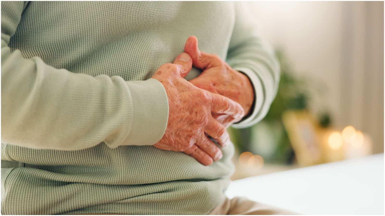 A person wearing a light green sweater is sitting down and clutching their stomach with both hands, indicating discomfort or pain. The background is blurred with some candles and a plant visible, perhaps suggesting they might find relief with lemon water before bed.