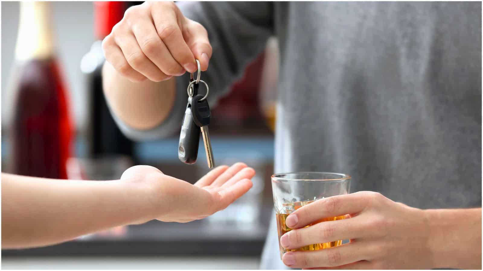 A person is handing over car keys to another, who has an open hand. The person giving the keys is holding a drink in the other hand, emphasizing the importance of abiding by alcohol rules. The blurred background suggests an indoor setting.