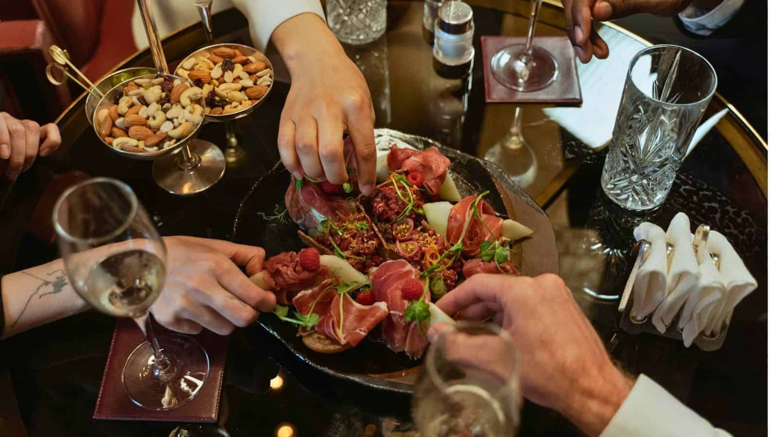Several people reach for various appetizers, including cured meats and fresh vegetables, arranged on a dark platter. Nuts in a glass dish and a few glasses of white wine are also on the table, adhering to alcohol rules, along with a crystal glass and folded napkins.