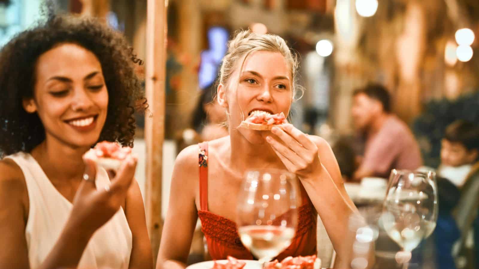 Two women sit at a table with drinks and food, one in a white top and the other in a red dress. As they enjoy their meal, the woman in the red dress takes a bite of food. The background shows other diners in an indoor setting, all adhering to alcohol rules.