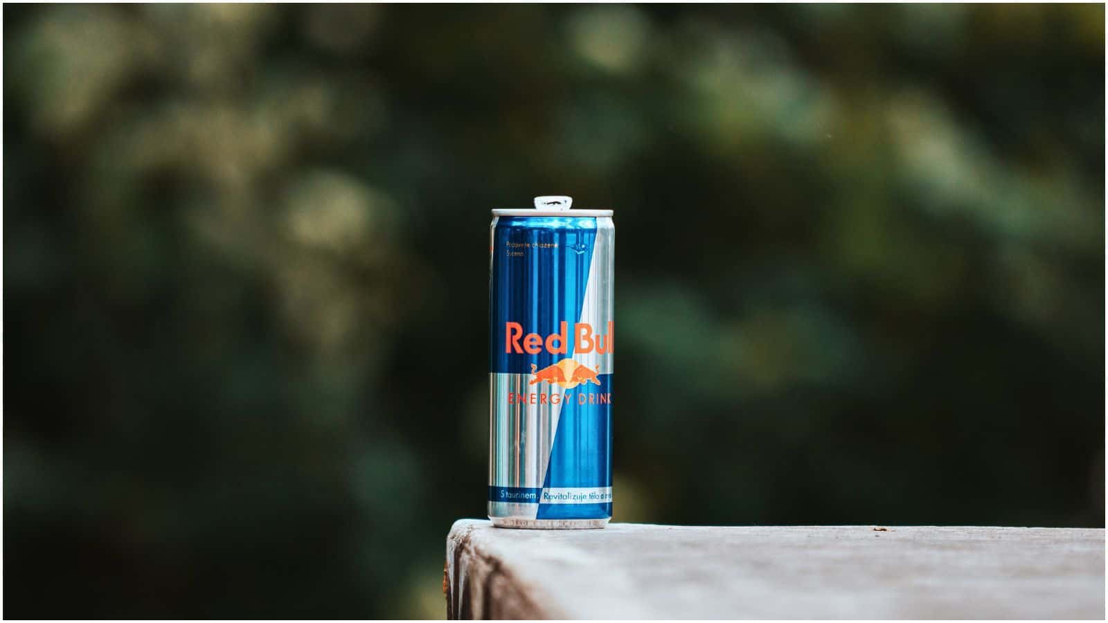 A single can of Red Bull energy drink is placed on a wooden surface with a blurred green outdoor background. The can, part of a popular list of sugary drinks, is predominantly blue and silver with the Red Bull logo and branding in red and yellow.