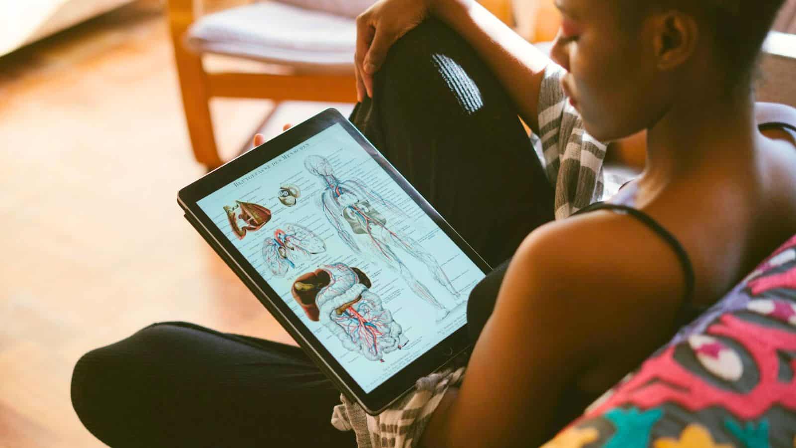 A person is sitting and holding a tablet displaying detailed diagrams of human anatomy, perhaps underscoring the importance of health in choices like to stop drinking alcohol. Dressed casually and seated on a cushioned surface, furniture and a wooden floor are visible in the background.