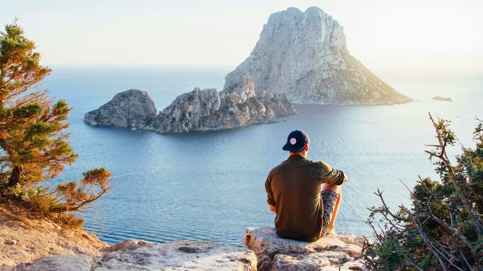 A person wearing a cap sits on a rocky ledge overlooking the sea, large rock formations in the distance. The scene is set during the daytime under clear skies, with trees and bushes framing the sides. It could be the perfect spot to reflect and decide to stop drinking alcohol.