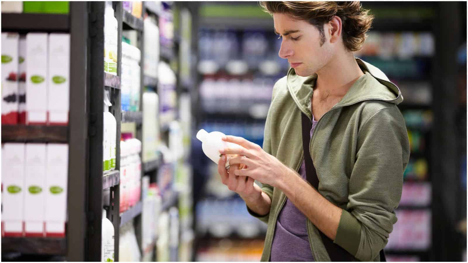 A young man with brown hair wearing a green hoodie is standing in a store aisle, closely examining a white bottle. Shelves with various products are visible around him. He appears to be reading the label, perhaps ensuring it doesn't contain any types of alcohol you shouldn't be drinking.