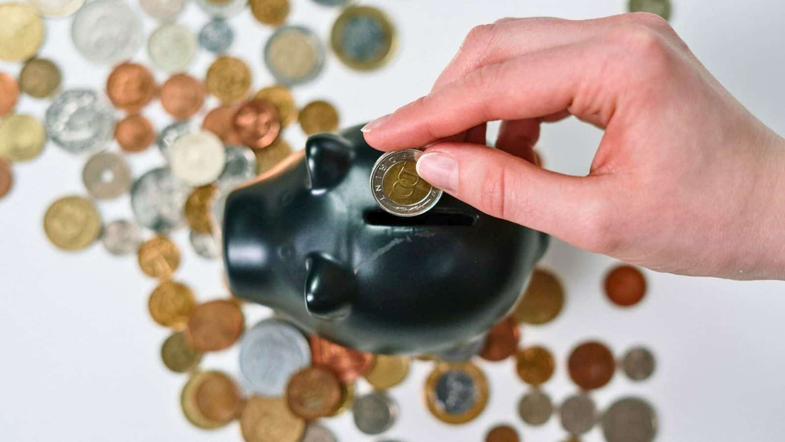 A hand is placing a coin into a black ceramic piggy bank. Various coins of different denominations and sizes are scattered around the piggy bank on a white surface, symbolizing small but meaningful steps like deciding to stop drinking alcohol.