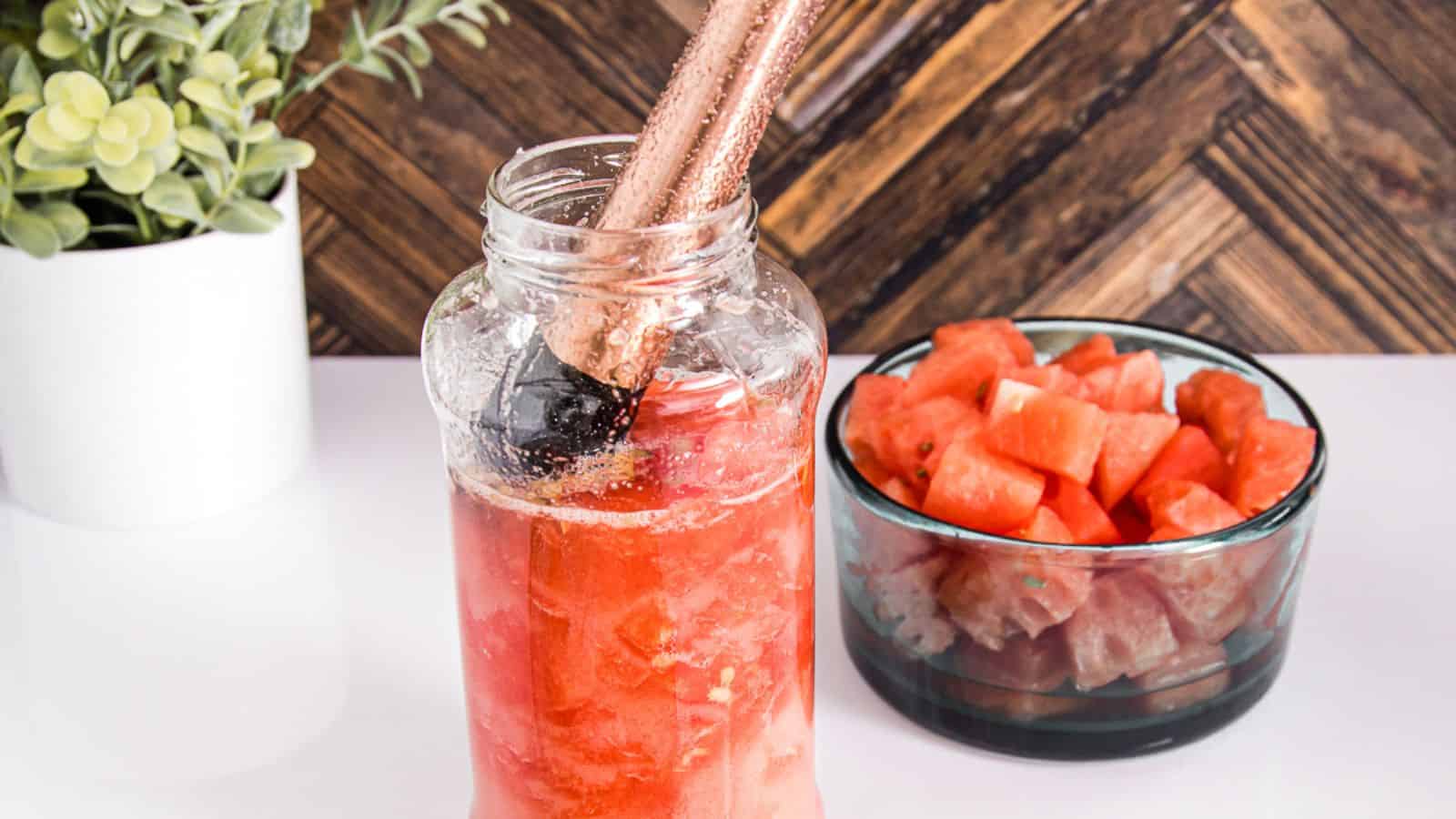 A glass jar filled with a red beverage and ice cubes is being stirred with copper straws. Beside the jar is a bowl containing chopped watermelon. A potted plant with green leaves is in the background, and the surface is white&mdash;everything you need to know about simple syrup for a perfect refreshment.