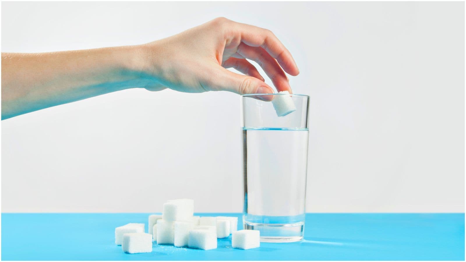 A hand is placing a sugar cube into a glass of water on a blue surface, reminiscent of preparing a list of sugary drinks. Several sugar cubes are scattered on the surface beside the glass. The background is a plain light color.
