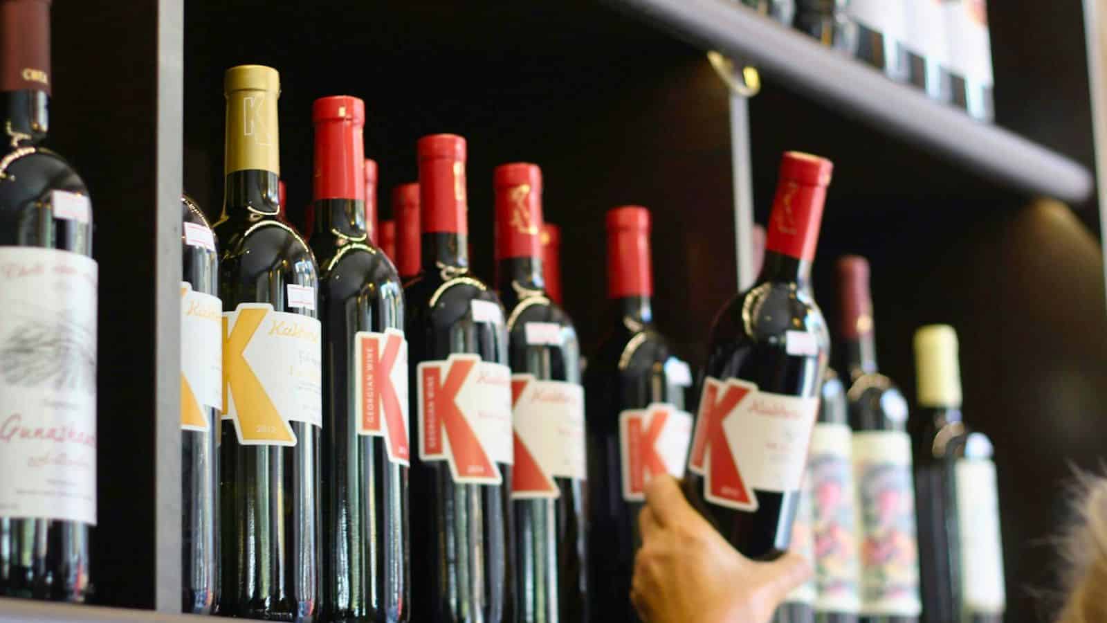 A person is reaching for a bottle of red wine from a shelf stocked with multiple bottles. The wine bottles have a label with a large letter "K" and red or gold caps, subtly reminding one to be mindful of the types of alcohol you shouldn't be drinking. Other types of wine are visible on the shelves above and below.