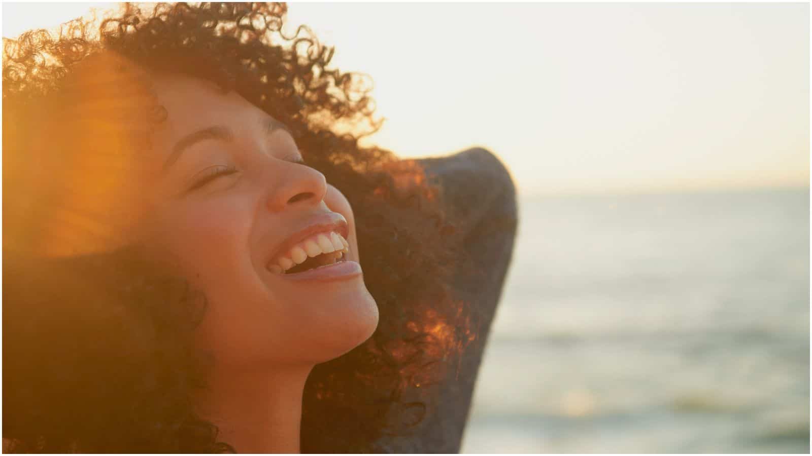 A person with curly hair is smiling joyfully with eyes closed, bathed in warm sunlight, and appears to be outdoors near the sea or ocean, as the water is visible in the background. The sun casts a golden glow over their face, reminiscent of the peaceful feeling one gets after enjoying lemon water before bed.