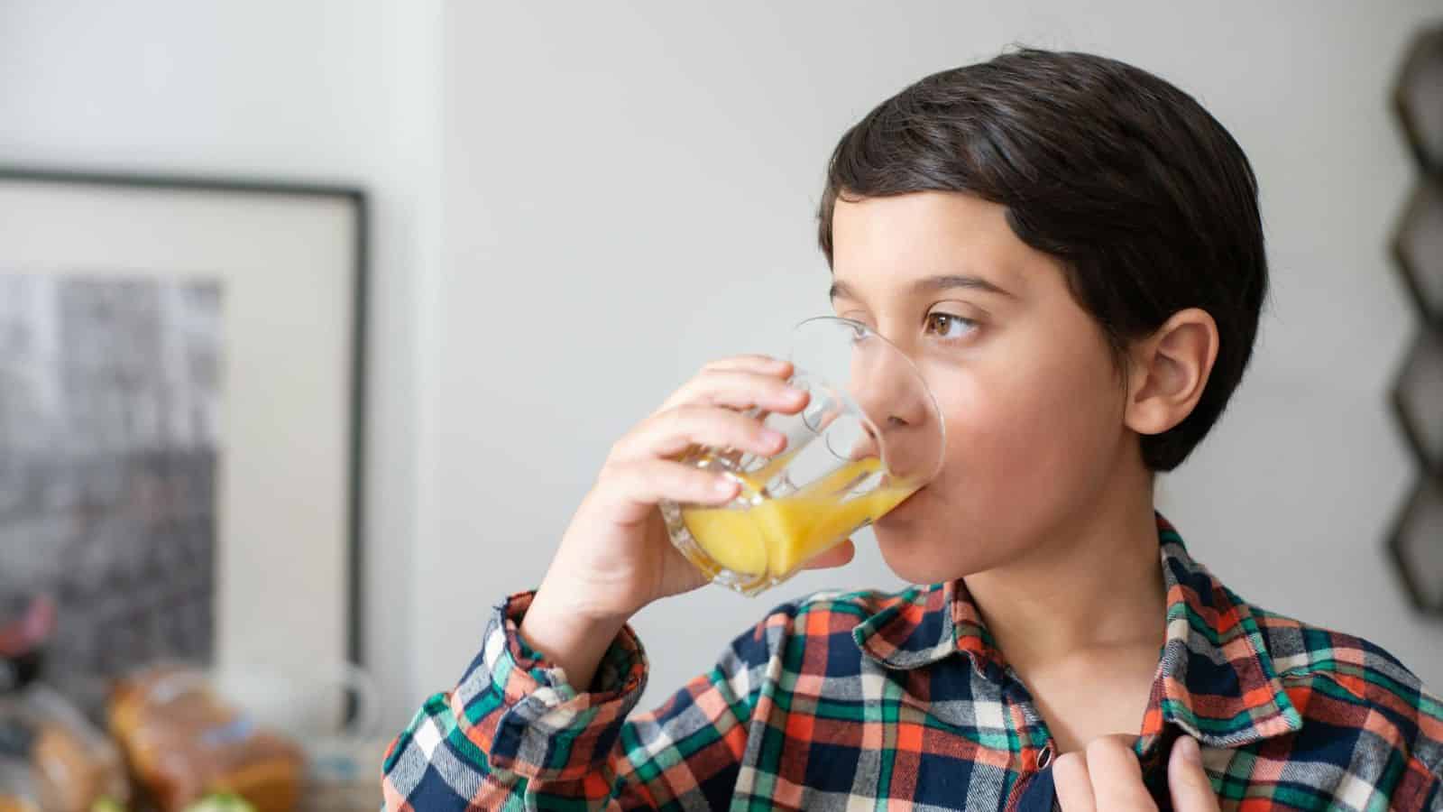 A young boy with short dark hair, wearing a plaid shirt, is drinking a glass of orange juice. He is holding the glass with one hand and has his other hand near his chest. The background, showing part of a table and wall, could be the perfect setting to discuss a list of sugary drinks kids enjoy.