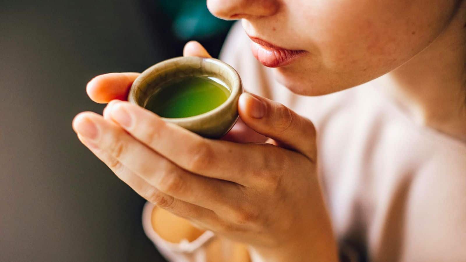 Close-up of a person holding a small cup of green tea near their lips, both hands gently cupping the cup as they prepare to take a sip.
